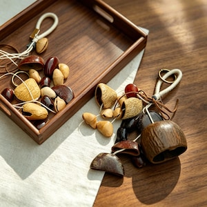 May include: A wooden tray filled with various natural, brown-toned decorative items. These include seed pods, beads, and bell-shaped ornaments, some strung with twine. A few items are arranged outside the tray on a wooden surface.