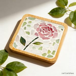 Puede incluir: Posavasos cuadrado con diseño de rosa en mosaico. El posavasos tiene un marco de madera y una superficie de mosaico con una rosa rosa, hojas verdes y detalles blancos y verdes. Mide 9cm x 9cm.