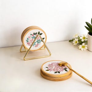 Puede incluir: Juego de posavasos de madera con dise&ntilde;o floral de mosaico en rosa, azul y verde. Los posavasos se exhiben en un soporte de metal dorado. Una cuchara dorada descansa sobre un posavasos. Accesorio decorativo.