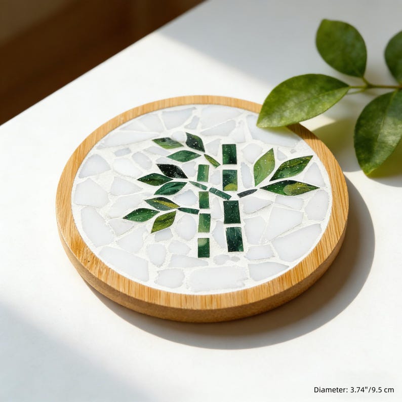 May include: Round coaster with a bamboo design. The coaster features a mosaic of white tiles with a green bamboo stalk and leaves. The coaster has a wooden rim. Diameter: 3.74 inches.