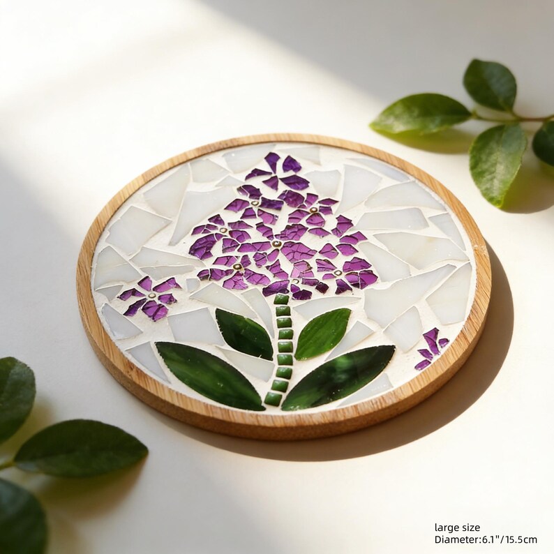 Puede incluir: Posavasos redondo de madera con dise&ntilde;o floral de mosaico. Presenta una flor morada con hojas verdes y fondo blanco. Di&aacute;metro: 15,5 cm.