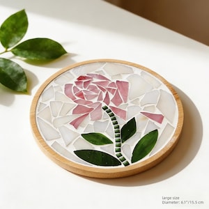 Puede incluir: Un posavasos redondo con un diseño de rosa en mosaico. La rosa está hecha de azulejos rosas y blancos, con hojas y tallo verdes. El posavasos tiene un marco de madera y mide 15,5 cm de diámetro.
