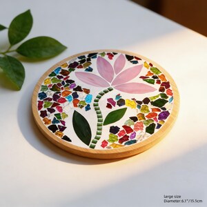 Puede incluir: Posavasos redondo de madera con un dise&ntilde;o de mosaico que presenta una flor rosa y piezas de vidrio de colores. El posavasos tiene un di&aacute;metro de 15,5 cm.