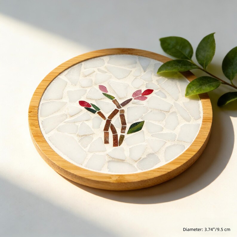 May include: Round coaster with a mosaic design featuring a stylized floral motif. The coaster has a light-colored wooden frame and a mosaic tile center with white, red, green, and brown elements. Diameter: 3.74 inches.