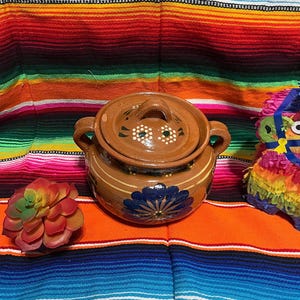 May include: A brown ceramic pot with a lid, featuring a blue floral design and white accents. The pot has two handles and is set against a colorful striped background, with a decorative succulent and a pinata.