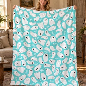 May include: A light blue fleece blanket featuring a repeating pattern of white ghosts with black eyes and mouths, interspersed with the word "Boo" printed in a simple white font across the entire surface of the fabric.