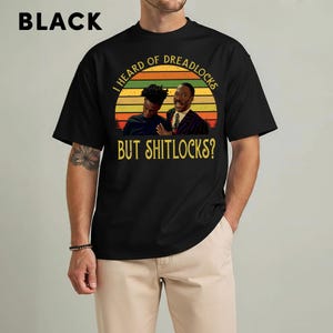May include: Black t-shirt with a graphic featuring two men and the text "I HEARD OF DREADLOCKS BUT SHITLOCKS?". The graphic is set against a sunset-style background with orange, yellow, and green stripes. The word "BLACK" is printed at the top.