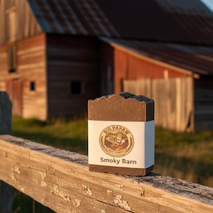 May include: A bar of brown soap with a white label that reads "Smoky Barn" and features a logo. The soap is resting on a weathered wooden fence, with a rustic barn in the background. The soap is likely handmade.