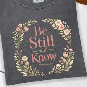 May include: A charcoal gray crew neck t-shirt featuring a floral wreath graphic surrounding the text "Be Still and Know" and "PSALM 46:10" in a soft pink font. The shirt has a Comfort Colors label at the inner neckline and a pepper color tag.