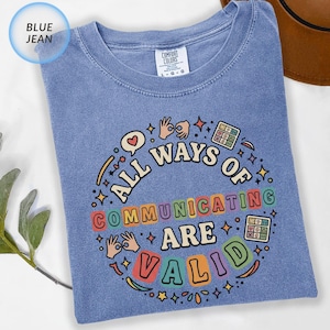 May include: A blue jean colored t-shirt with a graphic print that reads ALL WAYS OF COMMUNICATING ARE VALID. The design includes illustrations of hands signing, a speech bubble with a heart, and grid icons, all arranged in a circular pattern.