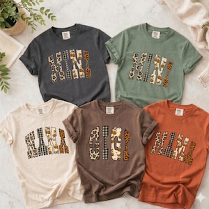 May include: Five short-sleeve t-shirts in charcoal, sage green, cream, brown, and burnt orange. Each shirt features a graphic print of a word in block letters filled with leopard, plaid, and pumpkin patterns. The words include MINI, NANA, GIGI, and MAMA.