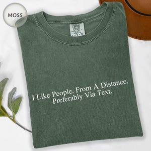 May include: Moss green t-shirt with the text "I Like People. From A Distance. Preferably Via Text." The shirt has a crew neck and short sleeves. The Comfort Colors brand tag is visible.
