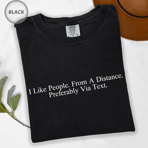 May include: Black t-shirt with the text "I Like People. From A Distance. Preferably Via Text." in white. The shirt is folded and has a Comfort Colors tag. A brown hat is partially visible in the background.