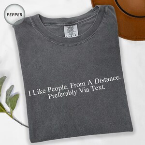 May include: Pepper-colored t-shirt with the text "I Like People. From A Distance. Preferably Via Text." printed in white. The shirt has a crew neck and short sleeves. The Comfort Colors brand tag is visible.