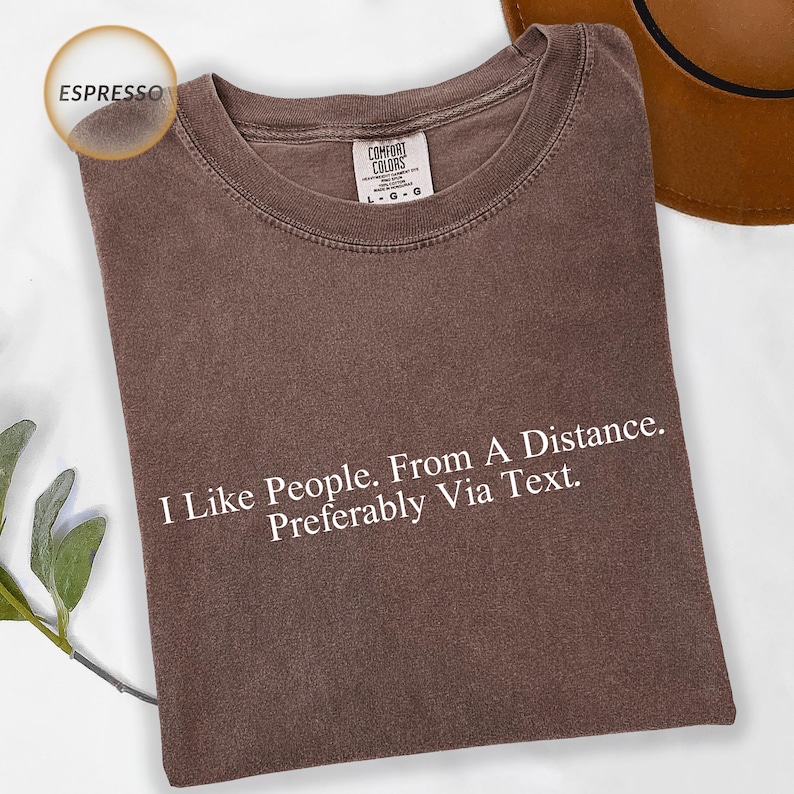 An espresso brown Comfort Colors t-shirt featuring clean white serif typography reading "I Like People. From A Distance. Preferably Via Text." Minimal text-only design with no illustrations, centered on the chest. Soft and understated aesthetic on a rich brown base. Designed for introverts and anyone with a dry, relatable sense of humor.
