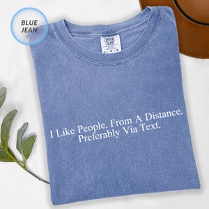 May include: A blue jean colored t-shirt with the text "I Like People. From A Distance. Preferably Via Text." printed in white. The shirt has a Comfort Colors tag.