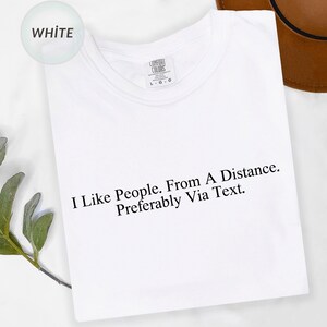 May include: White t-shirt with the text "I Like People. From A Distance. Preferably Via Text." in black. The shirt has a crew neck and a tag that says "Comfort Colors".