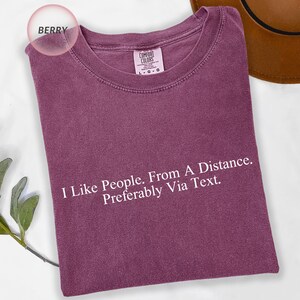 May include: Berry-colored Comfort Colors t-shirt with the text "I Like People. From A Distance. Preferably Via Text." The shirt has a crew neck and short sleeves. A brown hat is visible in the background.