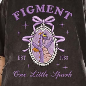 May include: Dark gray t-shirt with a purple embroidered design featuring the word "FIGMENT" above a purple bow and a framed image of a purple dragon. The text "EST 1983" and "One Little Spark" are also embroidered.