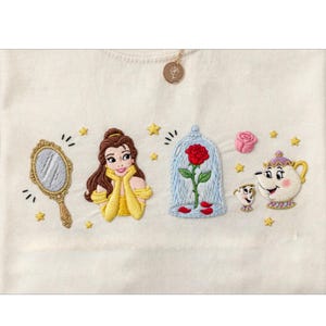 May include: Cream-colored embroidered design featuring Belle, a mirror, a rose in a glass case, a pink rose, and Mrs. Potts and Chip. Gold star accents and a gold pendant with a flower are also present.