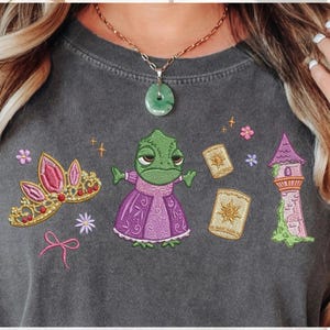 Puede incluir: Camiseta gris oscuro con elementos de dibujos animados bordados de un cuento de hadas. El diseño incluye un camaleón verde con un vestido morado, una corona rosa, linternas flotantes, una torre y detalles florales.