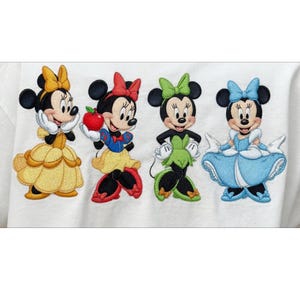 May include: Embroidered design featuring four Minnie Mouse characters dressed as Disney princesses. Each character wears a different outfit and bow, including yellow, red, green, and blue. The design is on a white background.