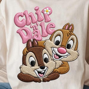 May include: Cream-colored t-shirt featuring an embroidered design of Chip and Dale, the Disney chipmunks. The text "Chip 'n Dale" is in pink, with a daisy accent. The chipmunks are brown, tan, and white, with large black eyes and pink noses.