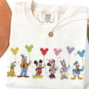 Könnte beinhalten: Cremefarbenes Comfort Colors T-Shirt mit Disney-Figuren, die Mickey Mouse-förmige Ballons halten. Zu den Figuren gehören Pluto, Goofy, Mickey, Minnie, Daisy und Donald Duck. Eine goldene Halskette liegt auf dem Shirt.