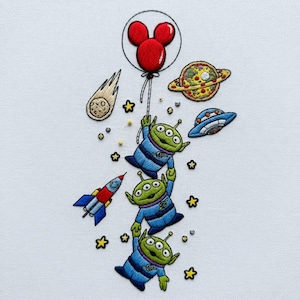 May include: Embroidered design featuring three green aliens in blue suits holding onto each other, ascending towards a red Mickey Mouse balloon. Includes a rocket, stars, a pizza planet, a comet, and a UFO on a white background.