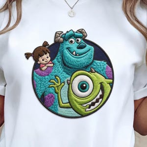 May include: White t-shirt featuring a colorful embroidered patch with characters from the animated film "Monsters, Inc.". The patch includes Sully, Mike Wazowski, and Boo, set against a dark blue circular background with purple accents.