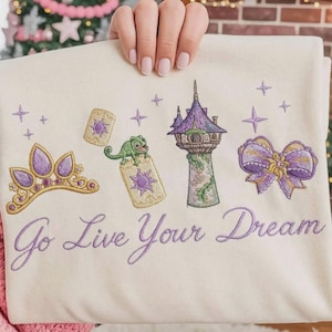 Könnte beinhalten: Cremefarbenes Sweatshirt mit gestickten lila und goldenen Details. Mit Krone, Turm, Schleife und dem Text "Go Live Your Dream". Enthält ein grünes Chamäleon und schwebende Laternen.