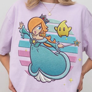 Puede incluir: Camiseta lila claro con un diseño bordado de un personaje de dibujos animados con cabello naranja, un vestido azul y una corona. El diseño incluye una estrella amarilla y una varita en forma de estrella. El fondo tiene rayas horizontales azules, rosas y blancas.