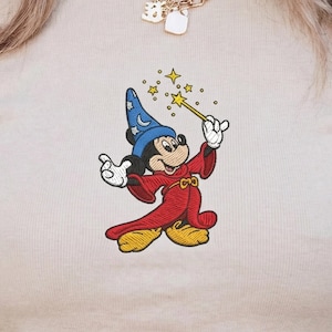 Könnte beinhalten: Gesticktes Design von Mickey Mouse als Zauberer, mit blauem Hut mit Sternen, rotem Umhang und gelben Schuhen. Er hält einen Zauberstab mit Stern, umgeben von kleineren Sternen. Das Design befindet sich auf hellem beigen Stoff.