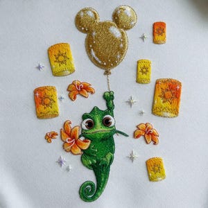 Puede incluir: Diseño bordado con un camaleón verde sosteniendo un globo dorado y flores naranjas. Alrededor del camaleón hay varias linternas amarillas y naranjas y acentos en forma de estrella. El diseño está sobre un fondo blanco.