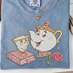 Puede incluir: Camiseta azul claro con un diseño bordado de la Sra. Potts y Chip de La Bella y la Bestia. El diseño incluye una rosa rosa, un libro y detalles dorados. También se ve un collar dorado con un colgante.