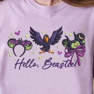 Op de afbeelding: Een lichtpaars T-shirt met geborduurde Halloween-ontwerpen. Het ontwerp omvat een vleermuisoren-hoofdband, een raaf met uitgespreide vleugels en een heksenhoed met een strik. De tekst "Hello, Beastie!" is ook op het T-shirt geborduurd.