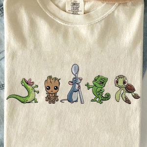 May include: Cream-colored t-shirt with embroidered cartoon characters: a green alligator, a brown Groot, a blue rat with a spoon, a green chameleon, and a turtle. The shirt has a Comfort Colors tag.