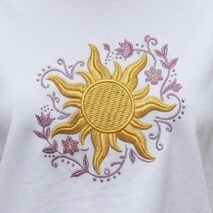 May include: White t-shirt with an embroidered golden sun design. The sun is encircled by purple floral and vine accents, creating a decorative and whimsical aesthetic. The embroidery adds texture and visual interest to the shirt, making it a unique piece.