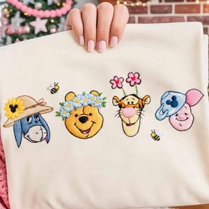 May include: Cream-coloured fabric with embroidered images of Winnie the Pooh characters: Eeyore in a straw hat, Winnie the Pooh with a flower crown, Tigger with flowers, and Piglet with a blue hat. Two bees are also embroidered.