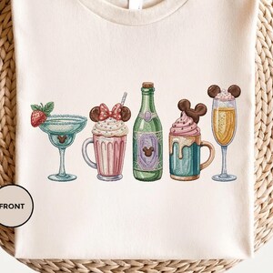May include: Cream-colored t-shirt featuring embroidered images of cocktails and drinks. The design includes a margarita, a milkshake, a champagne bottle, a hot chocolate, and a champagne flute, all with decorative elements.
