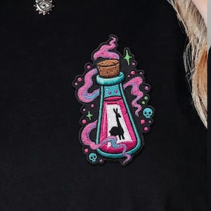 May include: Embroidered patch featuring a pink potion bottle with a black llama silhouette inside. The bottle has a teal base and a cork stopper. Pink and blue smoke-like swirls surround the bottle, with small skulls and stars.