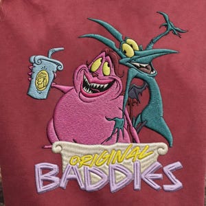 May include: Embroidered maroon sweatshirt featuring cartoon characters from the movie Hercules. The design includes the text "ORIGINAL BADDIES" in a stylized font. The characters are pink and teal, holding a drink.