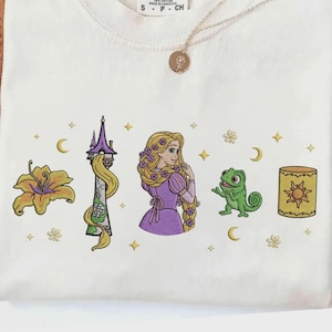 以下が含まれることがあります： クリーム色のTシャツ。黄色い花、長い金髪が垂れ下がる高い石の塔、紫色のドレスを着た長い金髪のキャラクター、緑色のトカゲ、黄色いランタンが刺繍されており、その周囲には星が散りばめられています。