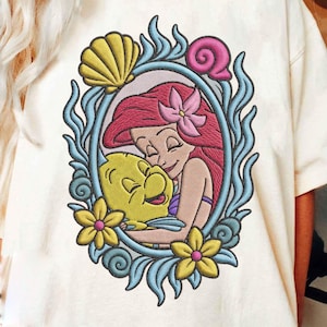 May include: Cream-coloured t-shirt with an embroidered design of a red-haired mermaid embracing a yellow fish. The design is framed by a light blue border with yellow flowers, a seashell, and a pink snail. The design is reminiscent of a popular animated film.