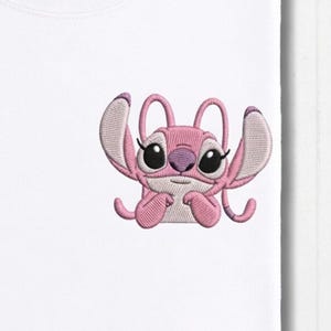 May include: White t-shirt featuring an embroidered pink cartoon character with large black eyes and a purple nose. The character has large ears and is in a playful pose. The design is centered on the shirt.