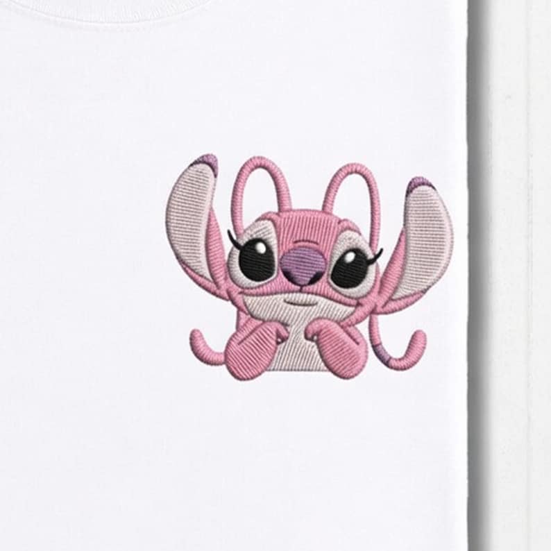 May include: White t-shirt featuring an embroidered pink cartoon character with large black eyes and a purple nose. The character has large ears and is in a playful pose. The design is centered on the shirt.
