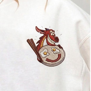 May include: White sweatshirt featuring an embroidered design of a red dragon holding a plate of fried eggs with chopsticks. The eggs are smiling. The dragon has a happy expression.