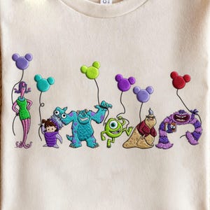May include: Cream-colored t-shirt featuring embroidered characters from the movie "Monsters, Inc." The design includes Mike Wazowski, Sully, Boo, and other characters, each holding a Mickey Mouse-shaped balloon in various colors. The design is centered on the shirt.