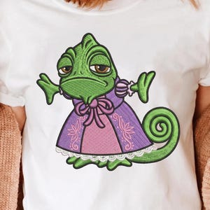 Peut inclure: T-shirt blanc avec une broderie d'un caméléon vert aux yeux marron, vêtu d'une robe violette et rose avec un nœud. Le caméléon a une queue enroulée et la broderie est sur le devant du t-shirt.