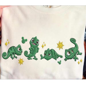 May include: Cream-colored sweatshirt featuring an embroidered design of four green chameleons in various poses. The design includes yellow suns, sparkles, and Mickey Mouse-shaped accents.